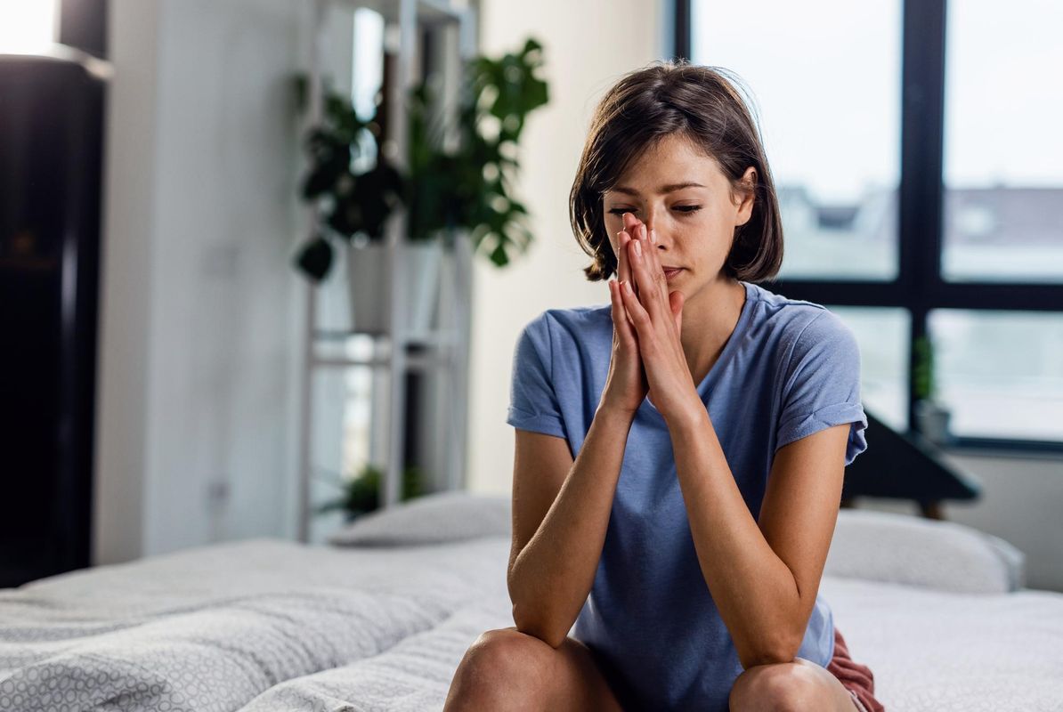 Uma mulher sentada na cama com os olhos fechados, com as mãos em frente ao rosto como se estivesse orando ilustra nosso artigo sobre: Medo: Três passos simples para lidar com essa emoção.