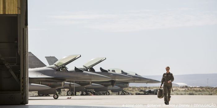 Brie Larson caracterizada como Carol Denvers caminha em direção ao hangar tendo ao fundo três F-16 com o cockpit aberto..