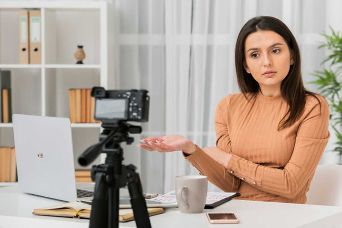 Imagem mostra uma câmera fotográfica filmando uma pessoa sentada numa mesa.