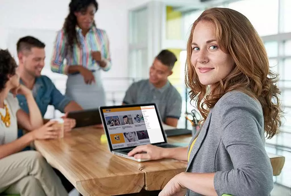 Uma mulher ruiva sorridente olha para a câmera enquanto está numa reunião de trabalho sobre Marketing digital. 