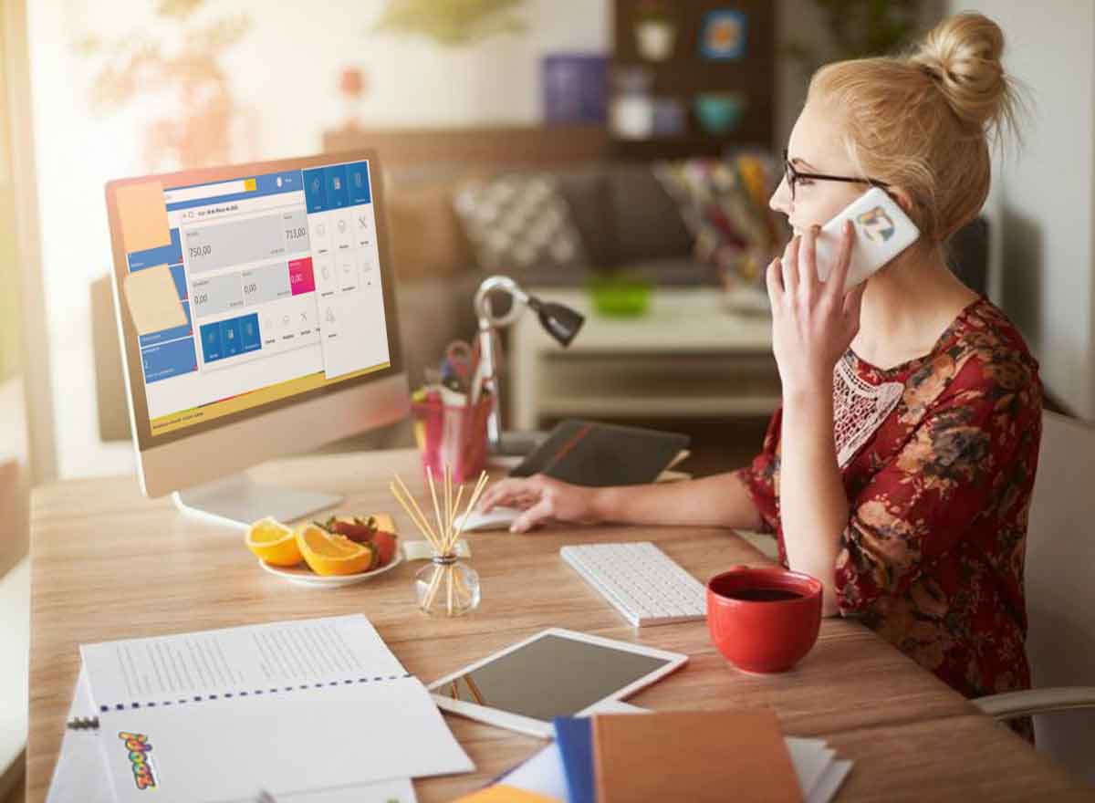 Uma mulher loira, jovem e usando óculos fala ao telefone enquanto olha para a tela do seu computador.