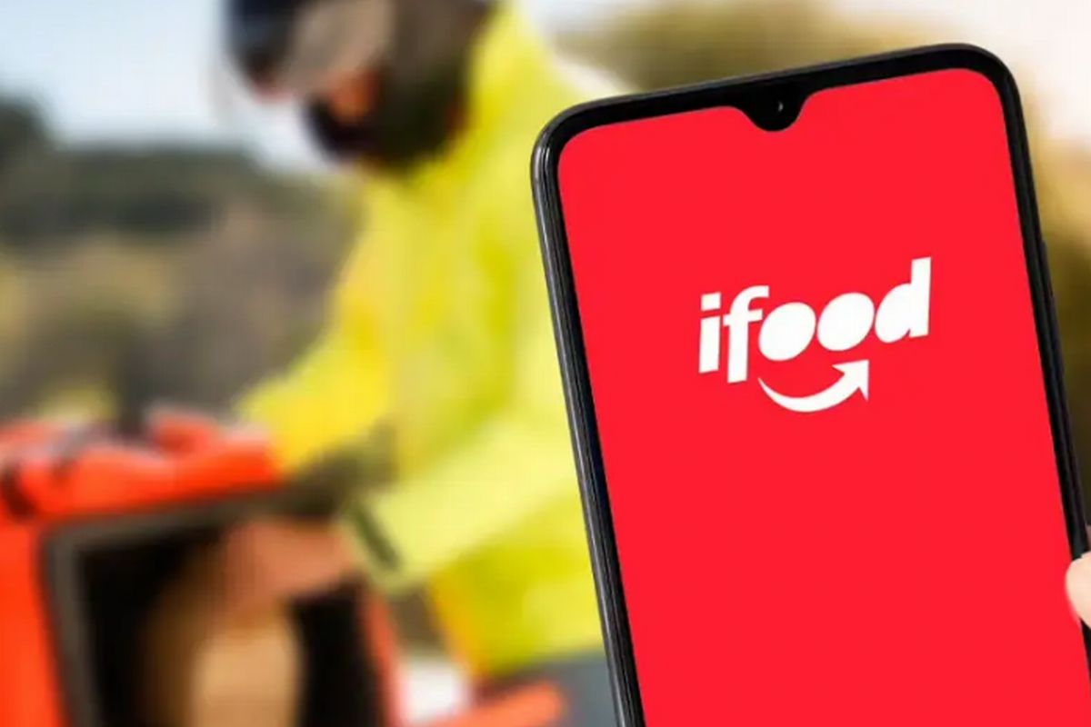 Uma pessoa segura um celular onde se pode ver a marca do ifood e ao fundo a imagem de um entregador, ilustra nosso artigo sobre: Telefone do iFood - Canais de atendimento da plataforma.  Foto: Divulgação/iFood