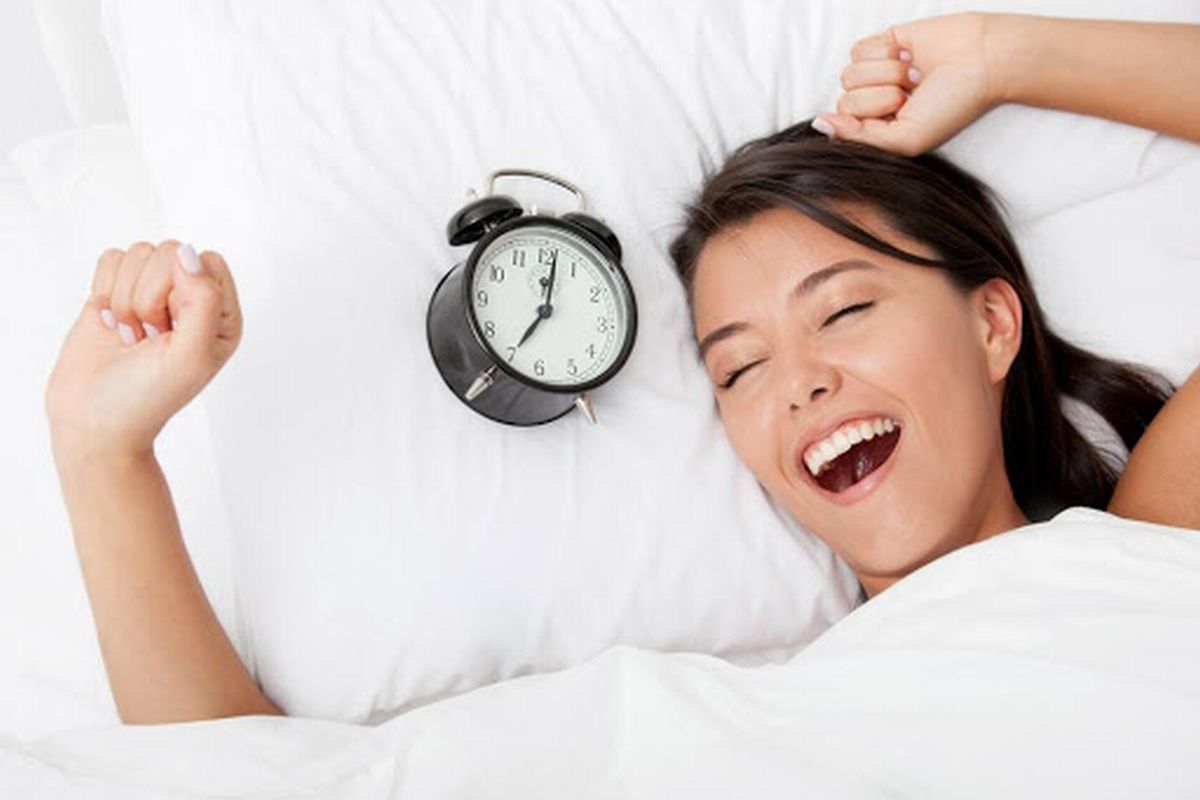 Uma mulher acordando feliz com o despertador ao lado ilustra nosso artigo sobre: Como dormir bem e acordar cheio de energia. (Foto: StockPhotos)